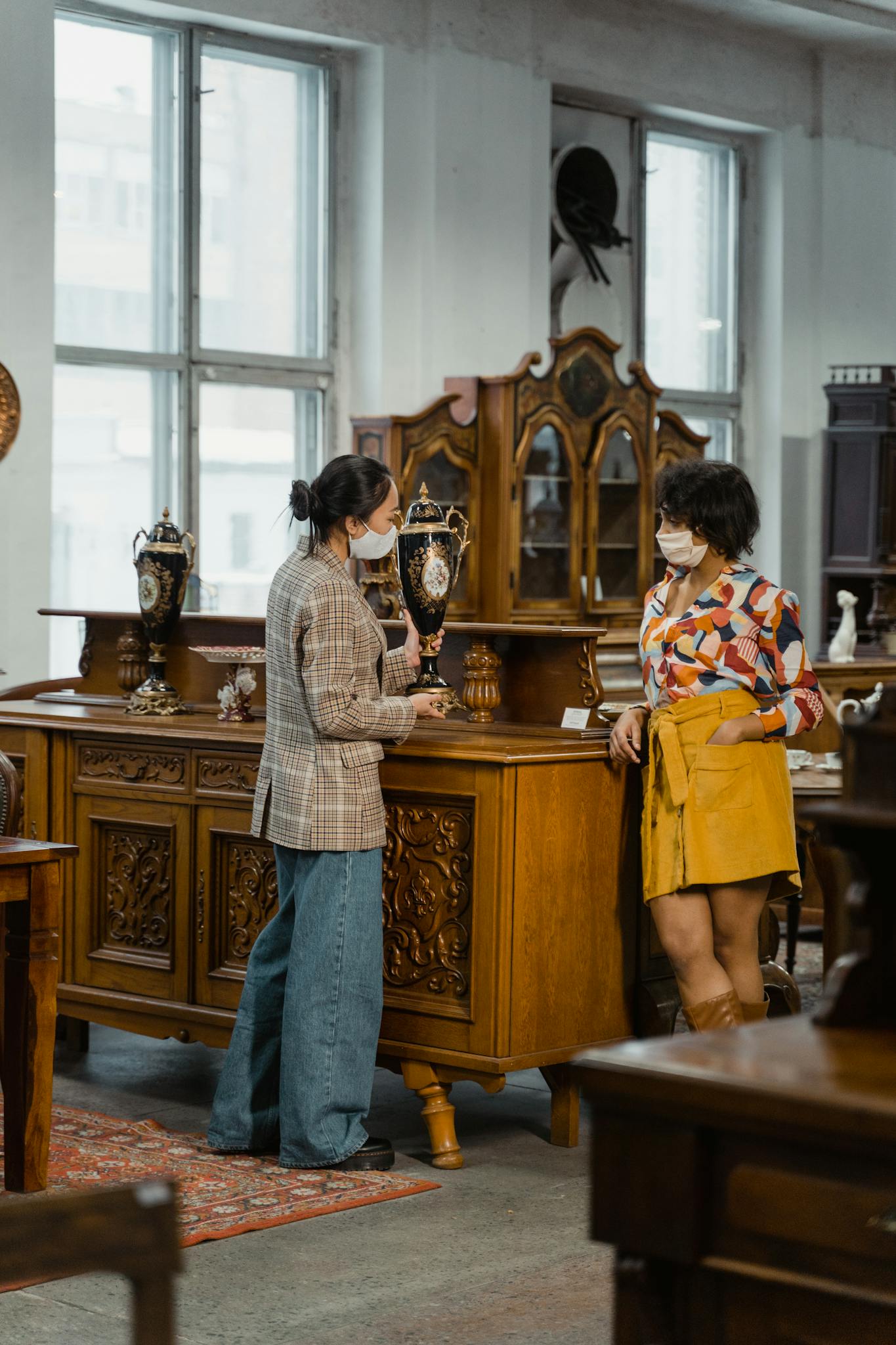 Two women wearing face masks browsing furniture in a vintage shop indoors.